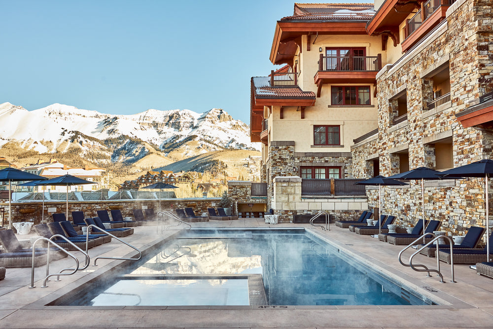 Heated outdoor pool at Madeline Hotel with lounge chairs and umbrellas, stone hotel facade, snow-capped mountains beyond