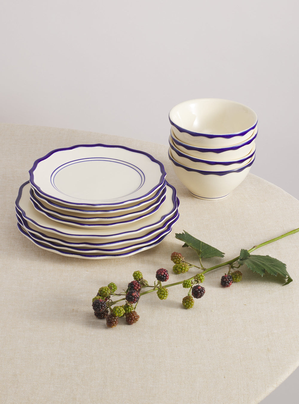Three-quarter view of Jane 12-piece dinnerware set showing cream stoneware with hand-painted cobalt scalloped rims on a beige linen tablecloth