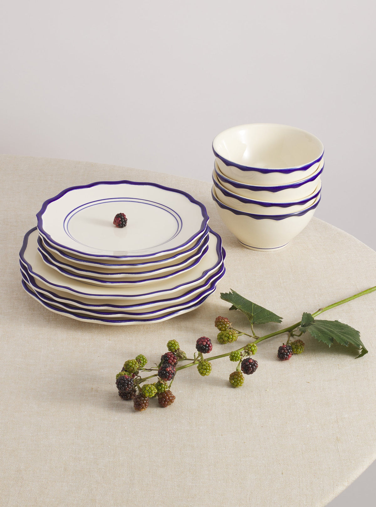 Front view of Jane 12-piece dinnerware set with stacked cereal bowls, cream glaze and cobalt-blue scalloped rims on linen