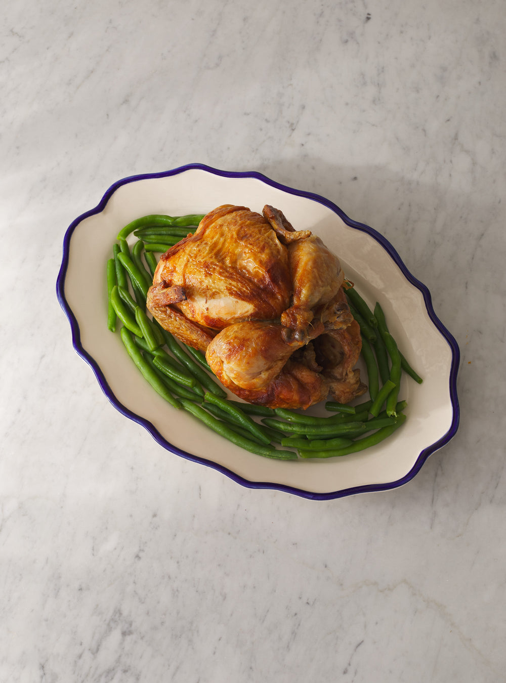 Overhead view of Jane Oval Platter showing scalloped edge and blue rim holding roasted chicken and green beans on marble