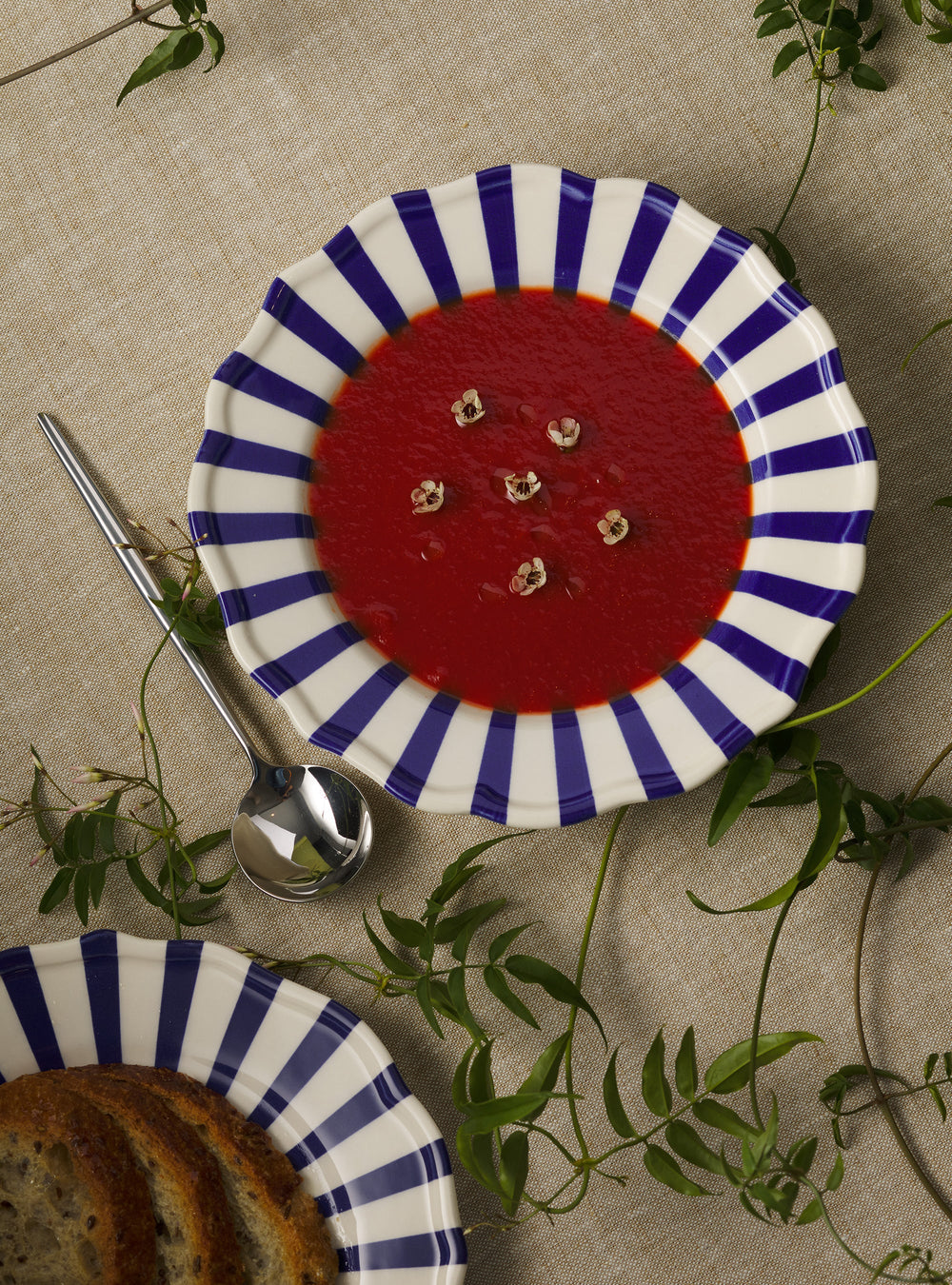 Top down view of Mafalda pasta bowl showing blue and white striped scalloped rim filled with red soup on linen background with spoon