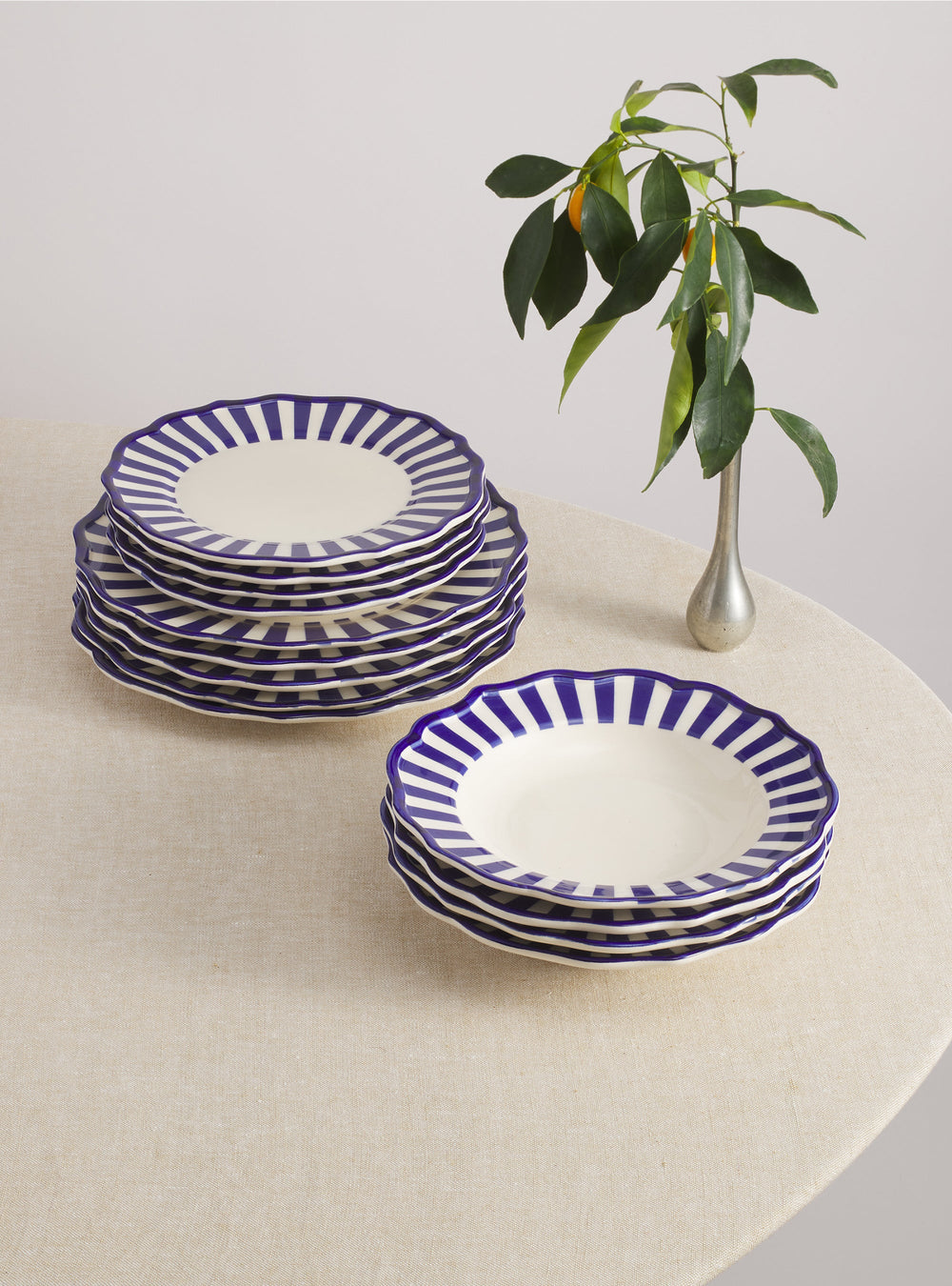 Three-quarter view of Riviera 12-piece dinnerware set showcasing blue striped scalloped rims on a linen-covered table with a small vase