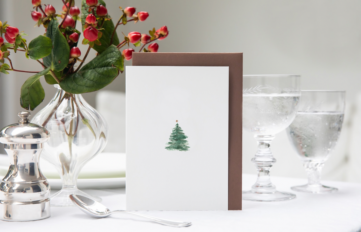Front view of Christmas Tree Cards, Set of 5 with a watercolor tree and star, brown nubuck envelope, and white table