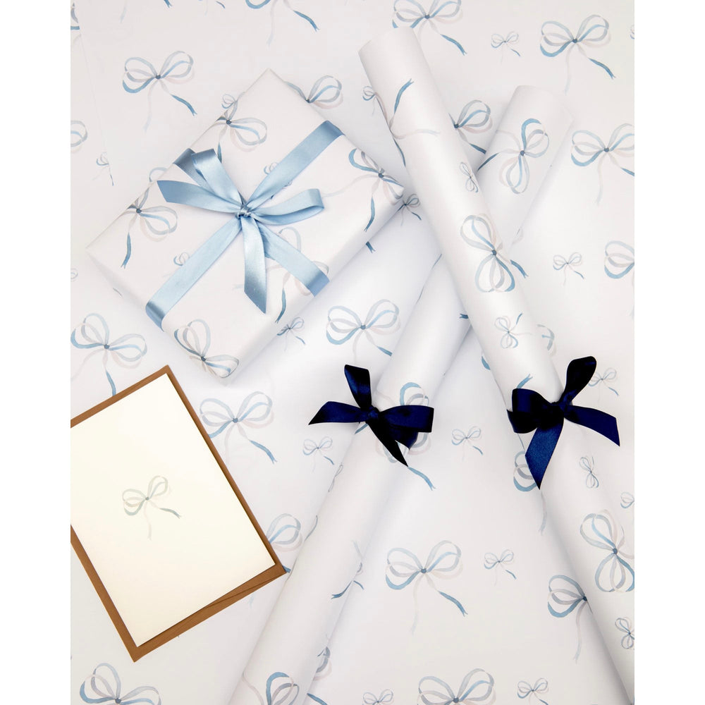 Flat lay of Lake Blue Bow wrapping paper sheets and rolls showcasing watercolor blue bow pattern with wrapped gift and coordinating ribbons on white background