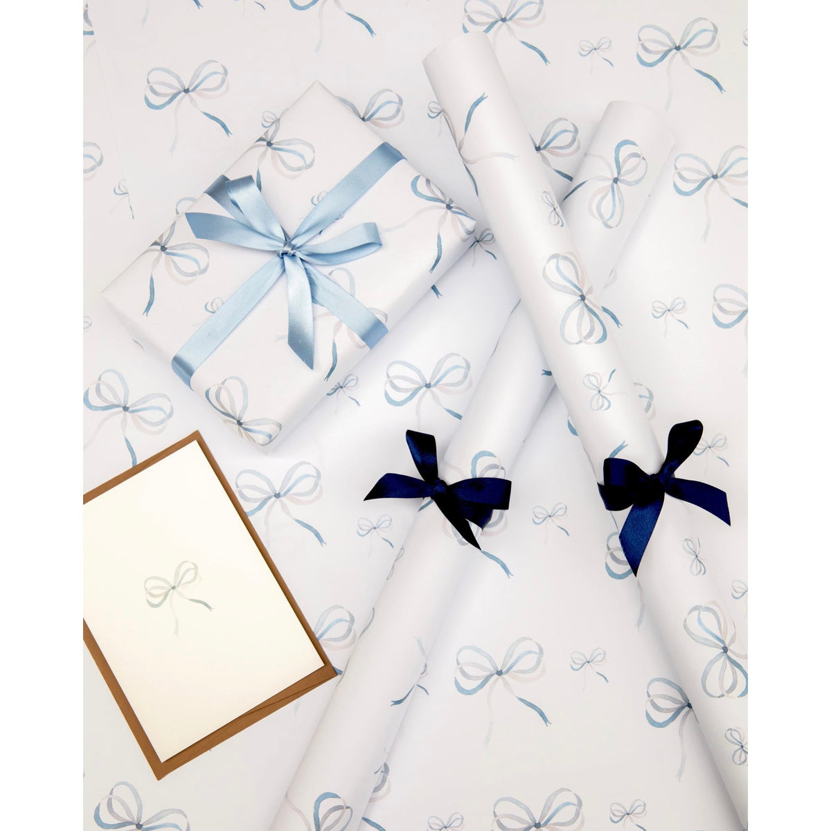 Flat lay of Lake Blue Bow wrapping paper sheets and rolls showcasing watercolor blue bow pattern with wrapped gift and coordinating ribbons on white background