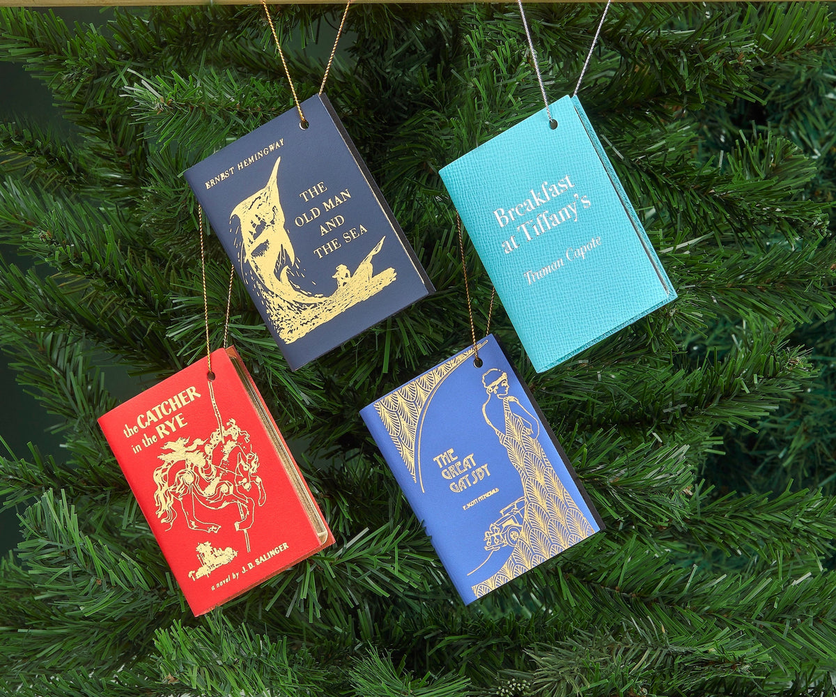 Four leather book ornaments hanging on an evergreen tree, featuring classic literary titles