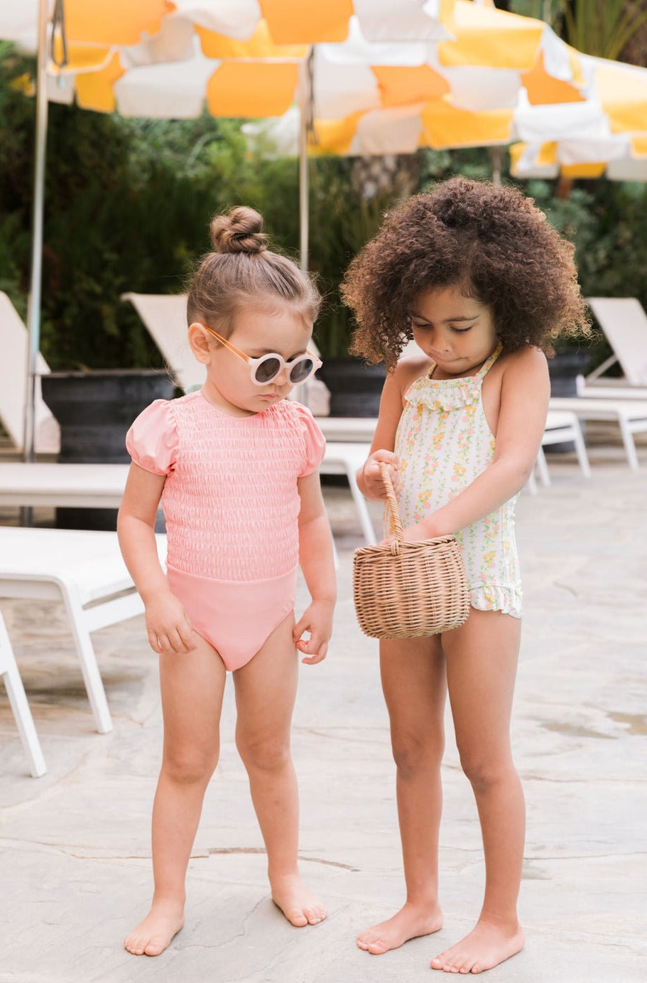 Two girls by a pool: one in a pink smocked swimsuit; the other in a citrus-print ruffle halter swimsuit holding a wicker basket