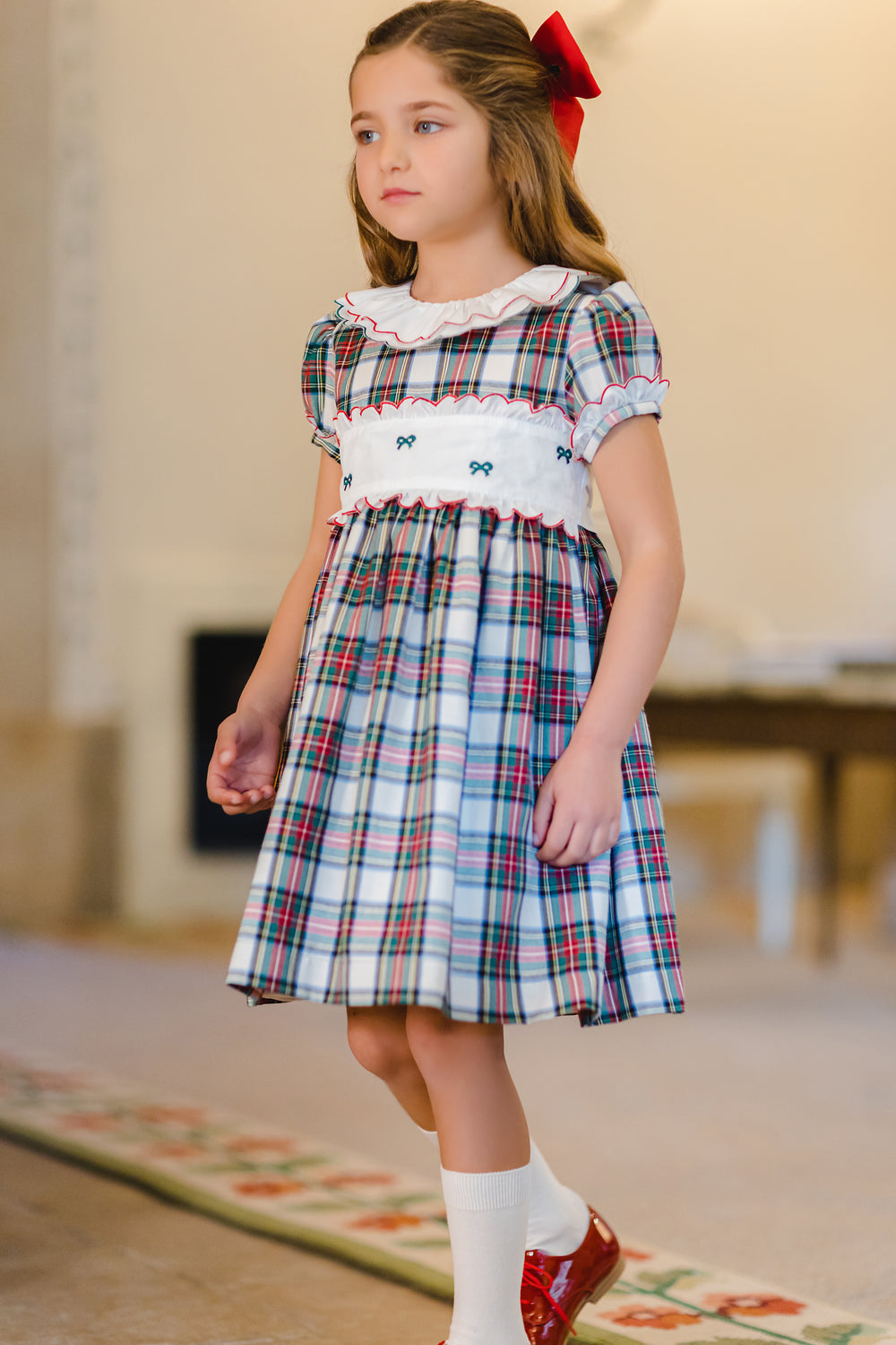 Three-quarter view of Mistletoe Dress on girl showing red-and-green tartan, white smocked waist and ruffled collar indoors
