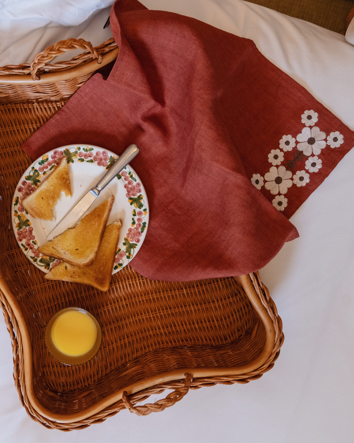 Brick red embroidered linen napkin draped over a wicker breakfast tray with a plate of toast and a glass of orange juice