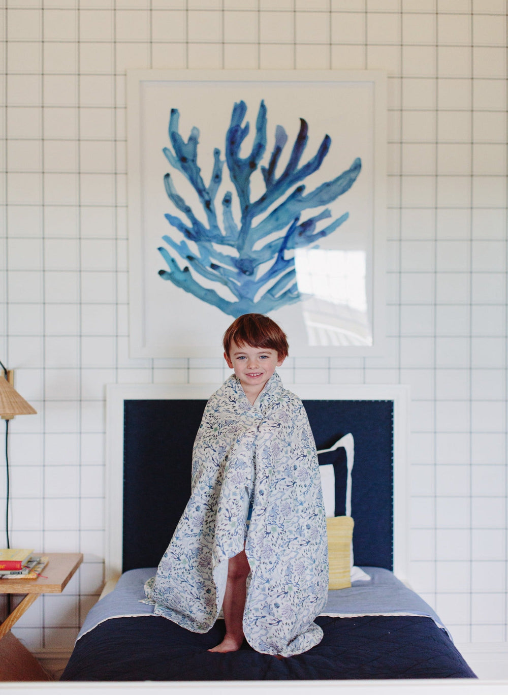 Young child wrapped in Coral Reef shawl blanket standing on a bed in a bright bedroom with blue coral artwork