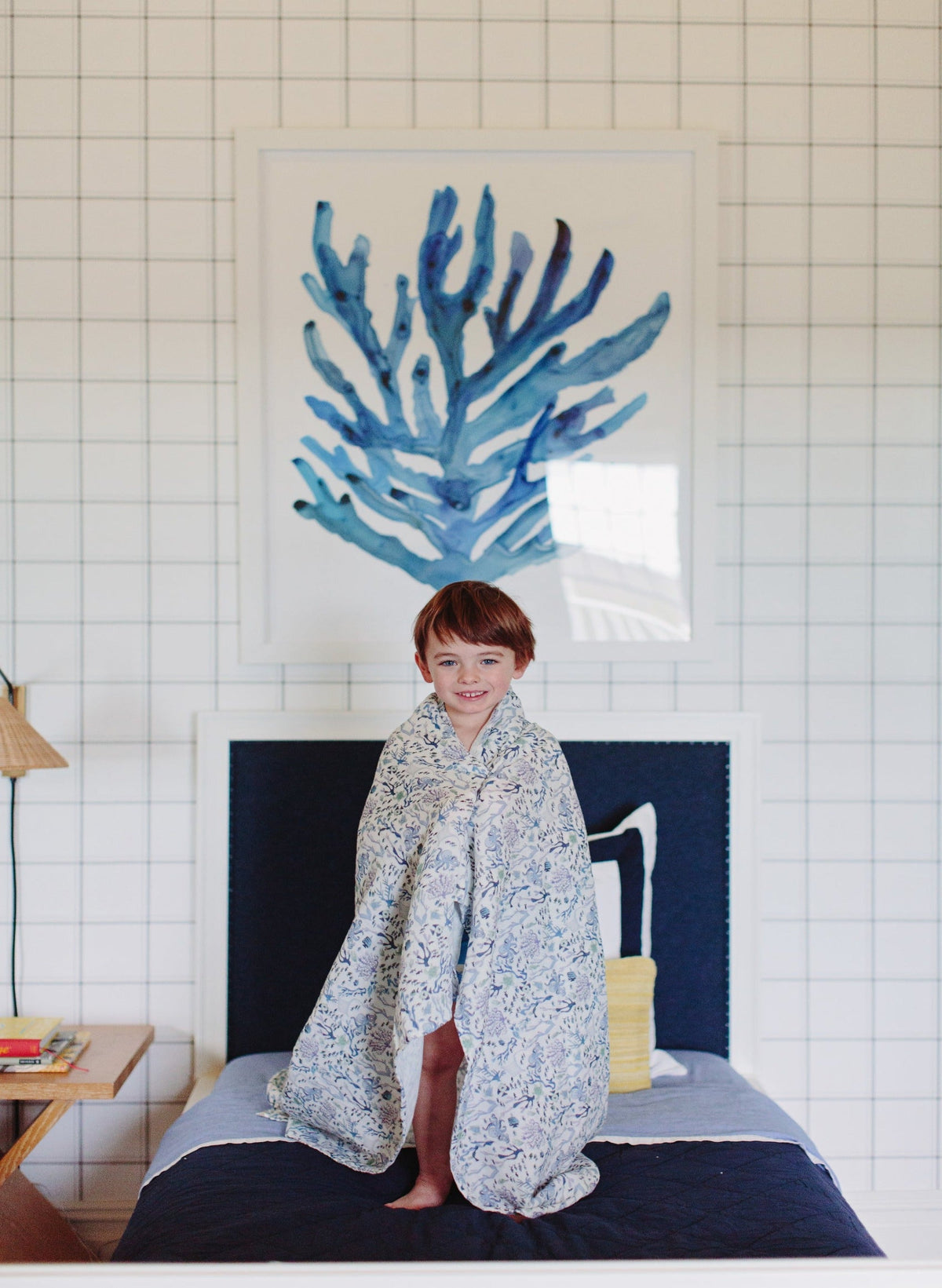 Young child wrapped in Coral Reef shawl blanket standing on a bed in a bright bedroom with blue coral artwork