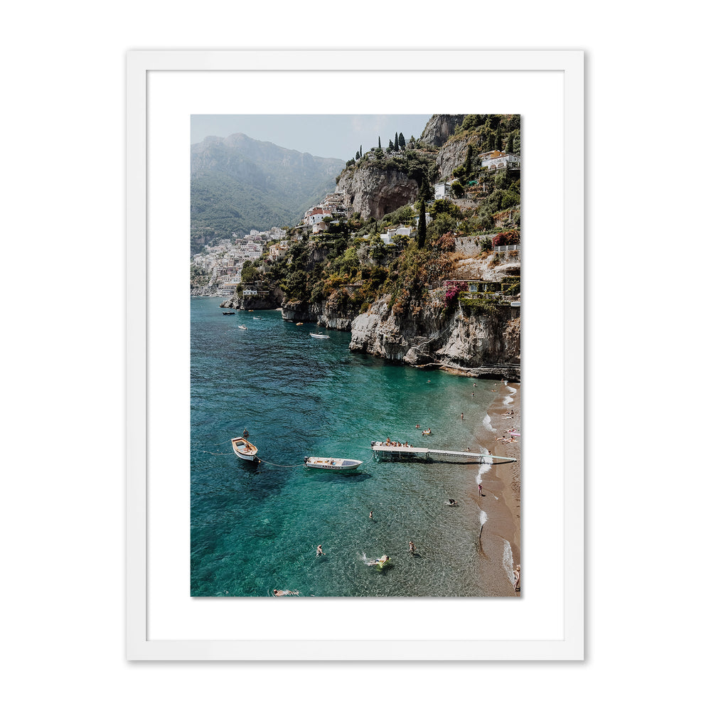 Front view of Bagni D’Arienzo II print showcasing a cliffside village with turquoise sea, boats, and beach, framed in white
