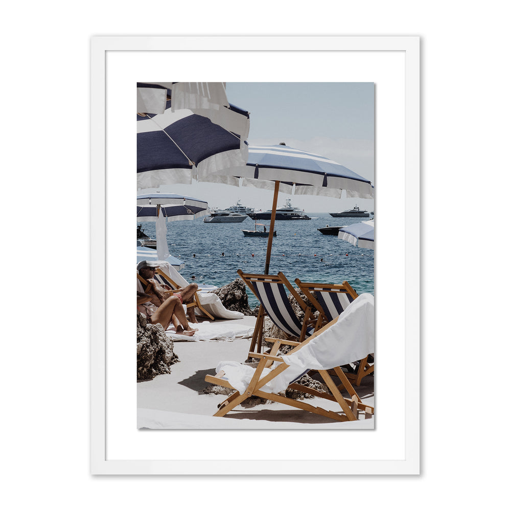 Front view of framed beach print with white mat showing blue and white striped umbrellas, wooden deck chairs, and yachts on the sea