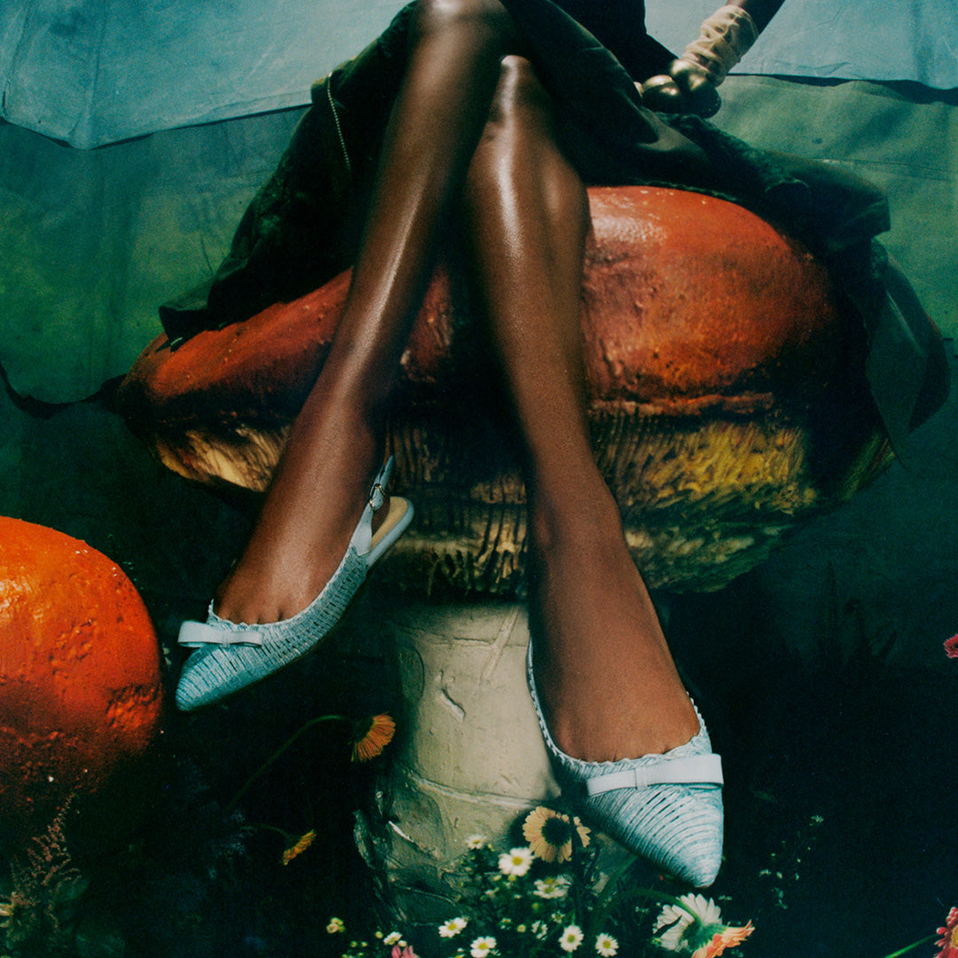 Three-quarter view of Natalie Sling in Seafoam Raffia showcasing pointed toes and asymmetrical bows on a model among mushrooms