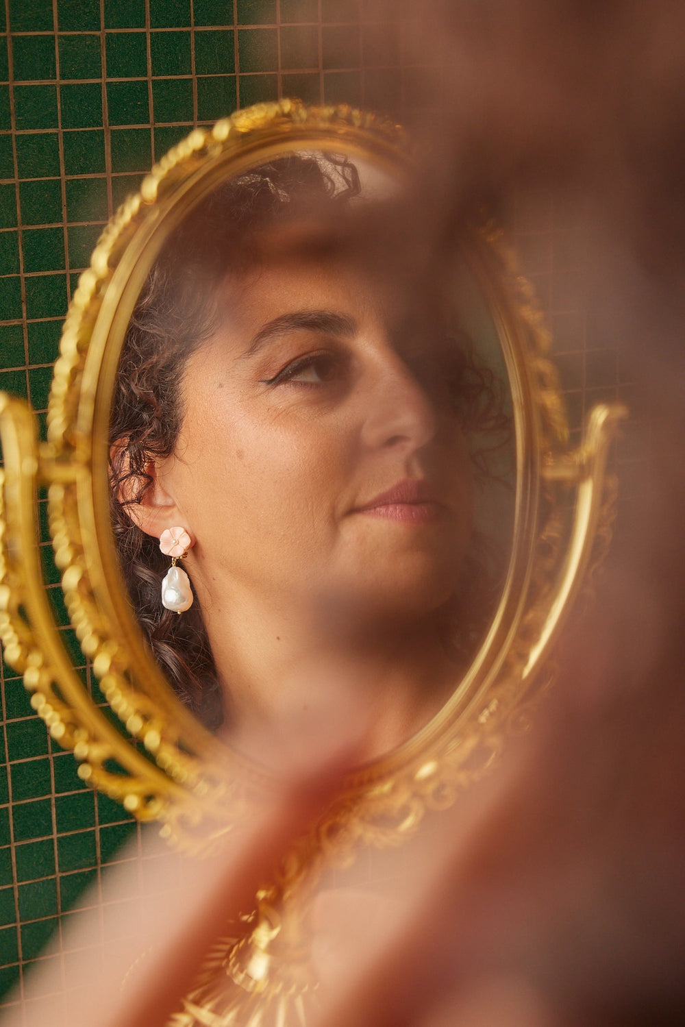 Woman looking into an ornate gold mirror wearing pink pearl drop earrings against a green tiled wall
