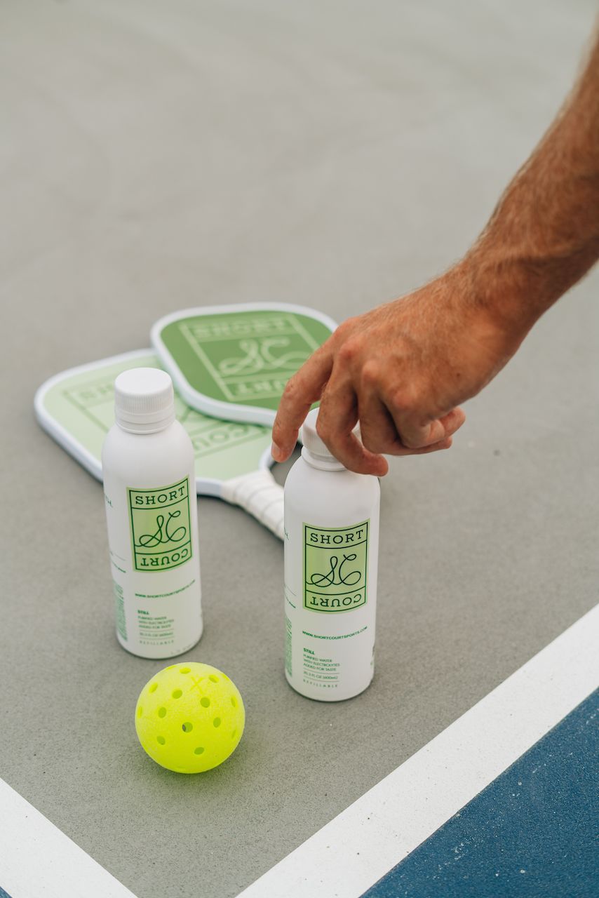 Overhead view of Doubles bundle with two paddles, two branded white bottles, and a neon yellow pickleball on a court