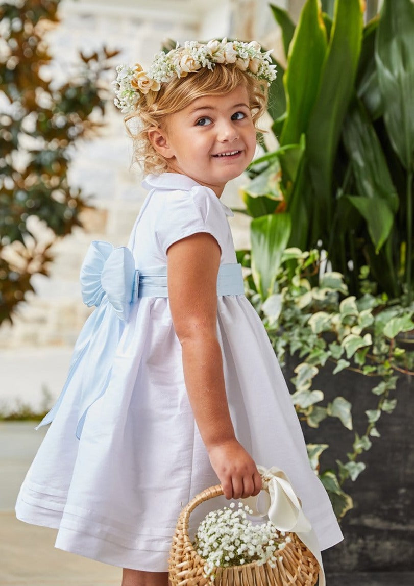 Side view of Peter Pan formal dress in Special Occasion White on a child, showing Peter Pan collar, cap sleeves and light blue bow sash against a garden background