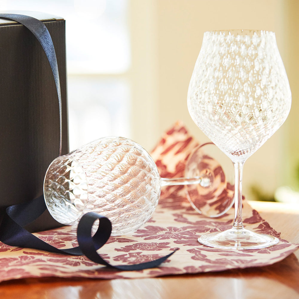 Three-quarter view of Phoebe Clear universal wine glasses, set of 2 showcasing diamond-optic mouth-blown crystal with gift box and navy ribbon on patterned cloth