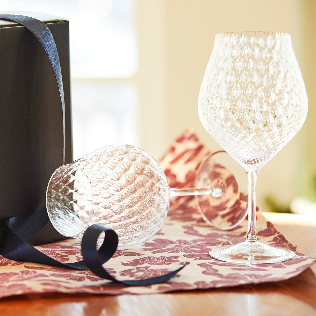 Three-quarter view of Phoebe Clear universal wine glasses, set of 2 showcasing diamond-optic mouth-blown crystal with gift box and navy ribbon on patterned cloth
