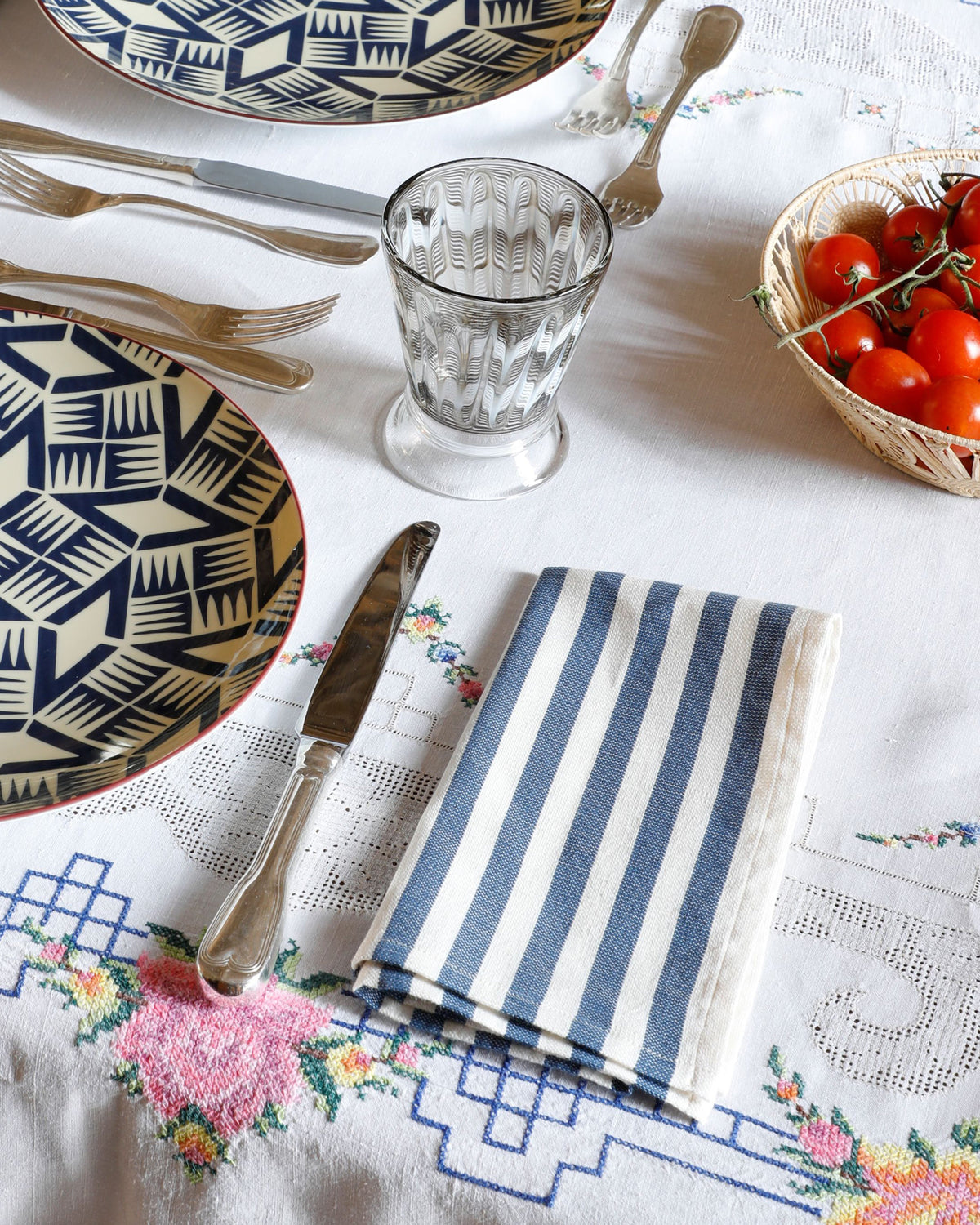 Light blue striped napkins arranged on a table with patterned plates, silverware, glassware, and tomatoes