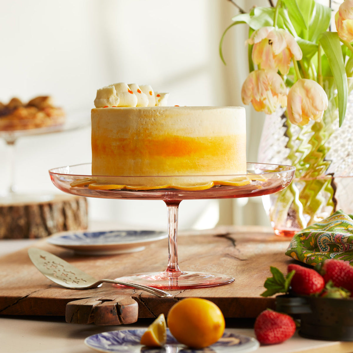 A Celia rose pink crystal glass cake stand from Caskata holds a bright yellow cake on a Spring table.