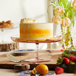 A Celia rose pink crystal glass cake stand from Caskata holds a bright yellow cake on a Spring table.