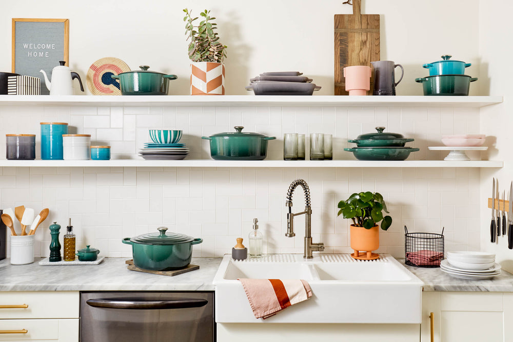 Artichaut Le Creuset Signature Braiser on kitchen countertop with matching cookware on open shelves above a farmhouse sink