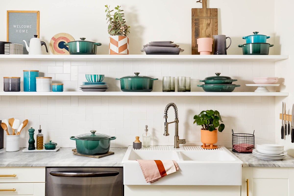 Artichaut Le Creuset Signature Braiser on kitchen countertop with matching cookware on open shelves above a farmhouse sink