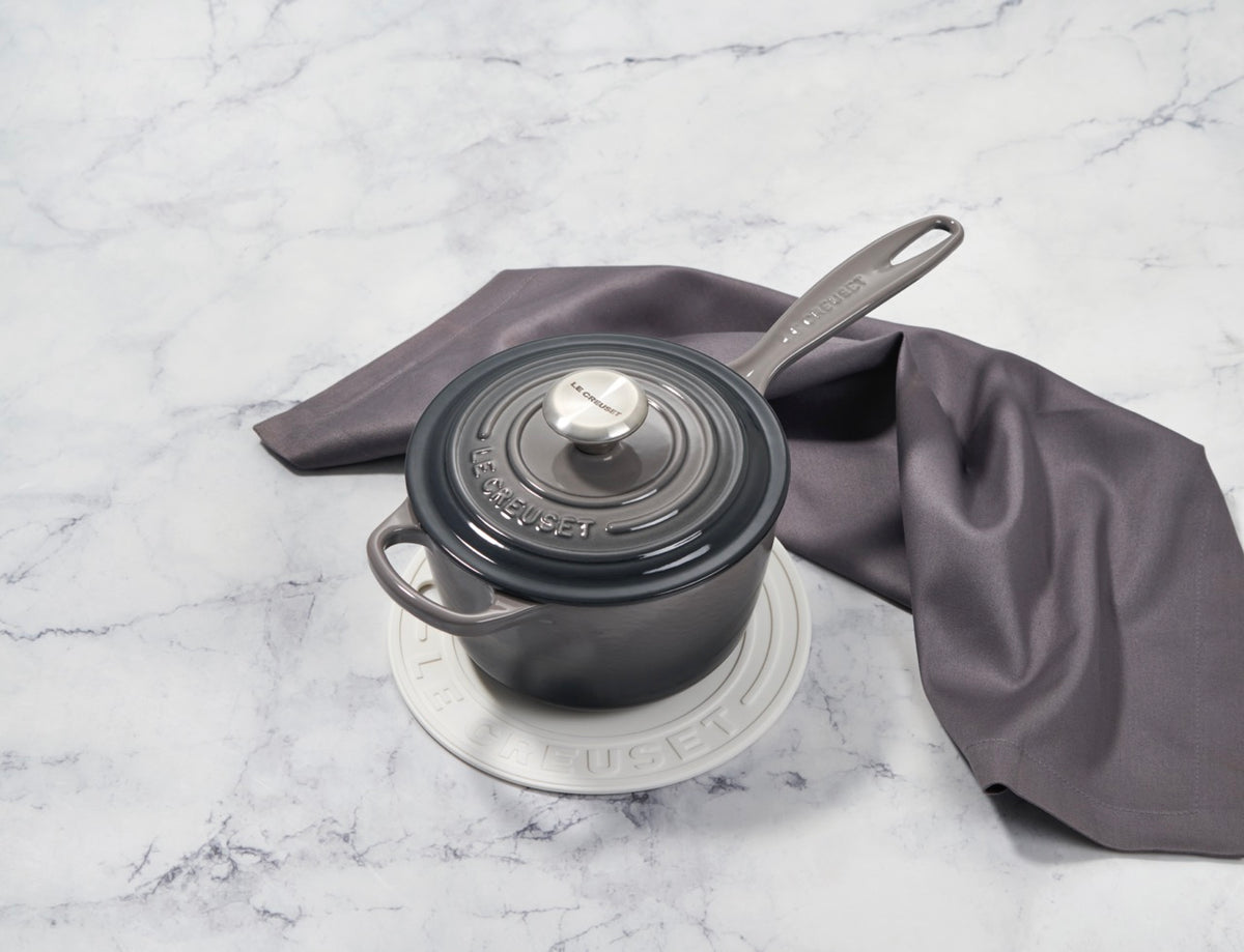 Top-down view of a white Le Creuset silicone trivet under a small covered gray saucepan on a marble surface with folded gray linen