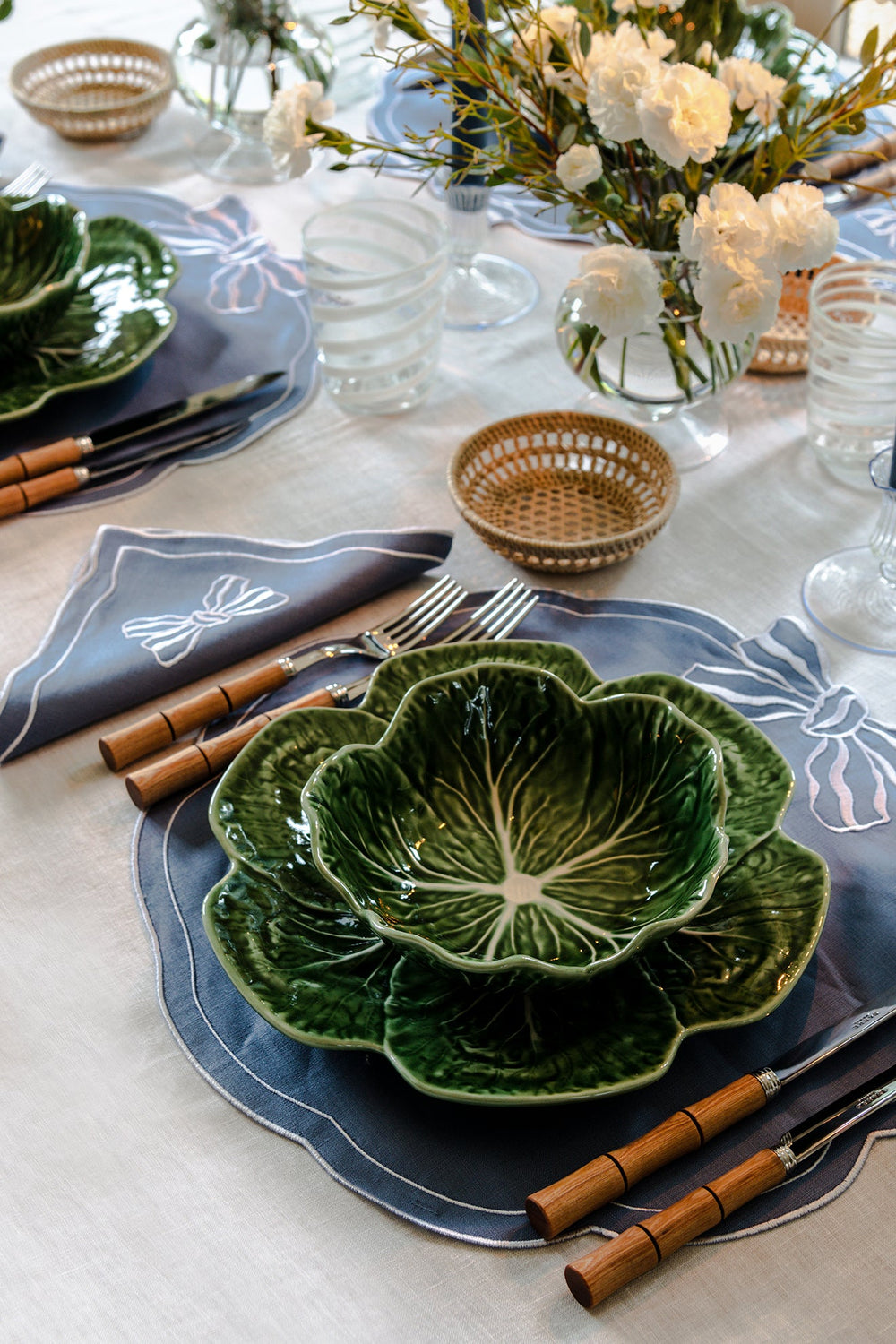Ivory hemstitch linen tablecloth with green cabbage bowls, wood-handled flatware, blue placemats, and white flowers