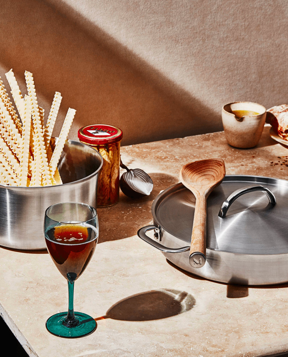 Angled overhead view of The Copper Set showcasing stainless-steel finish pans with lid and wooden spoon on a stone countertop