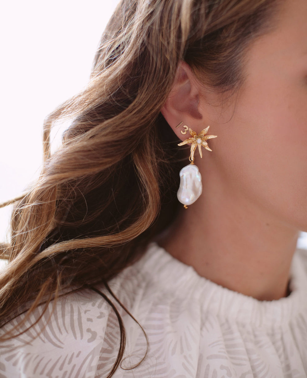 Close-up of Rehearsal Earrings showcasing textured gold starburst stud and dangling irregular baroque pearl on a model against a light background