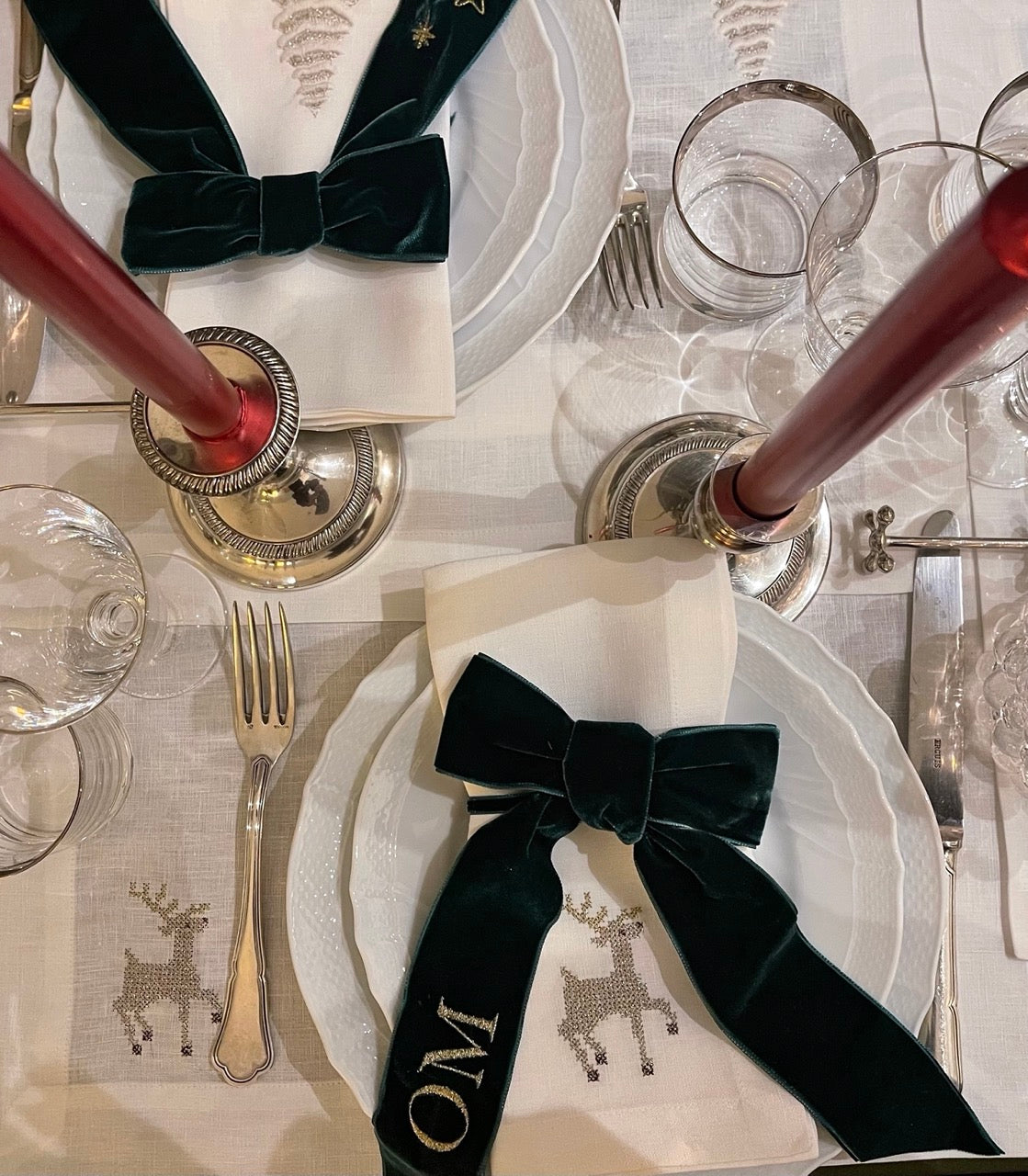 Holiday table with linen placemat embroidered with reindeer, white plates, green velvet bow napkin, and red taper candles