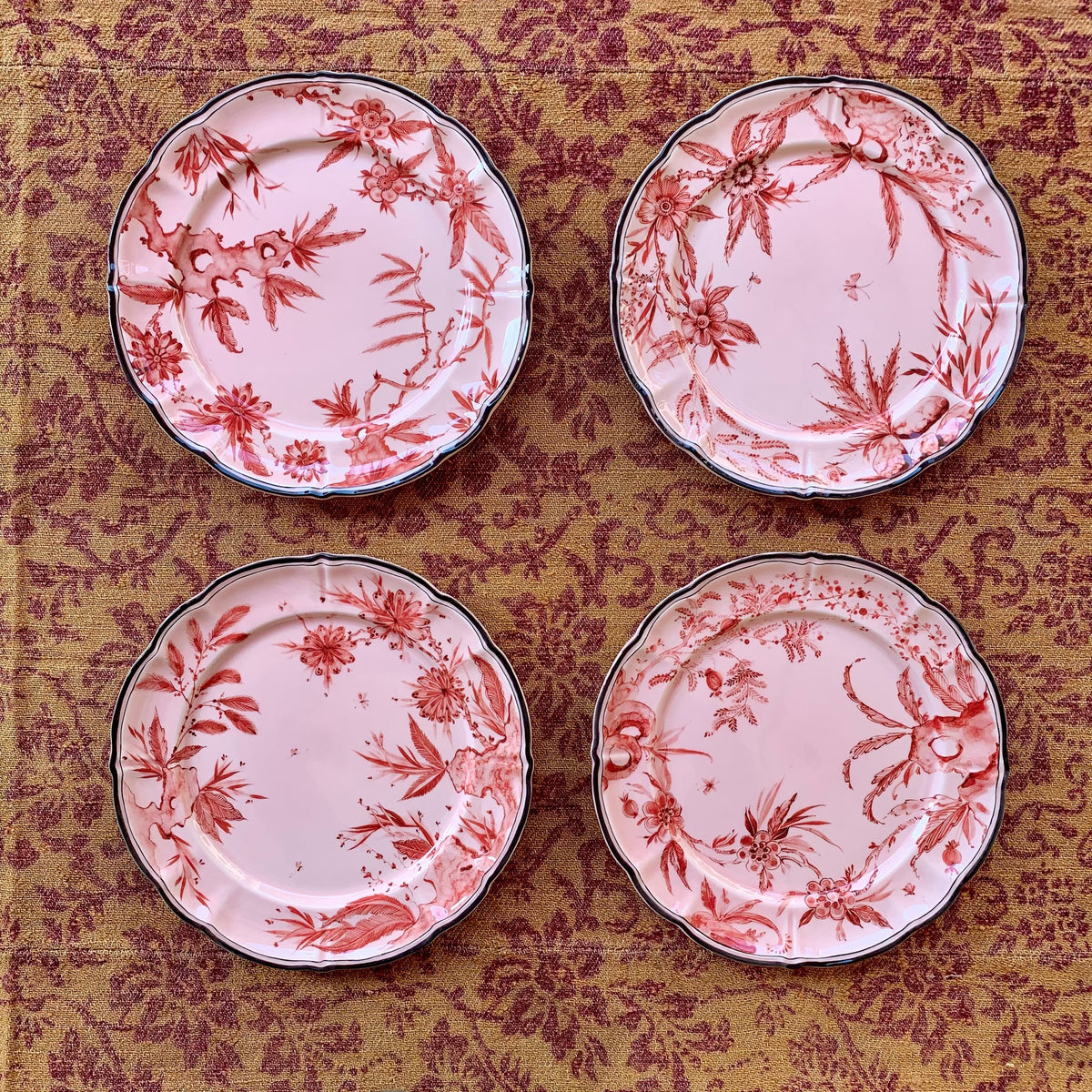 Top-down view of four Rocaille Pink dessert plates showing red floral transferware pattern and scalloped rims on woven fabric