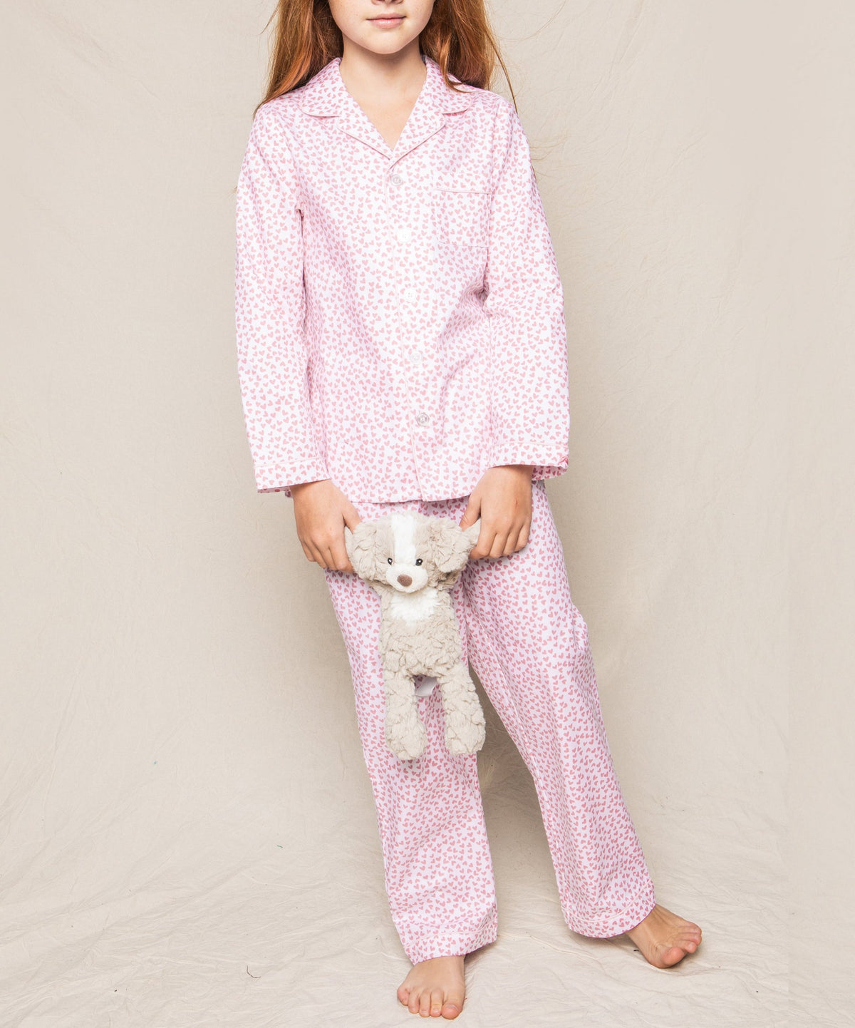 Front view of Sweethearts pajama set on child model showing pink heart-print long-sleeve button-up top and matching pants on beige background