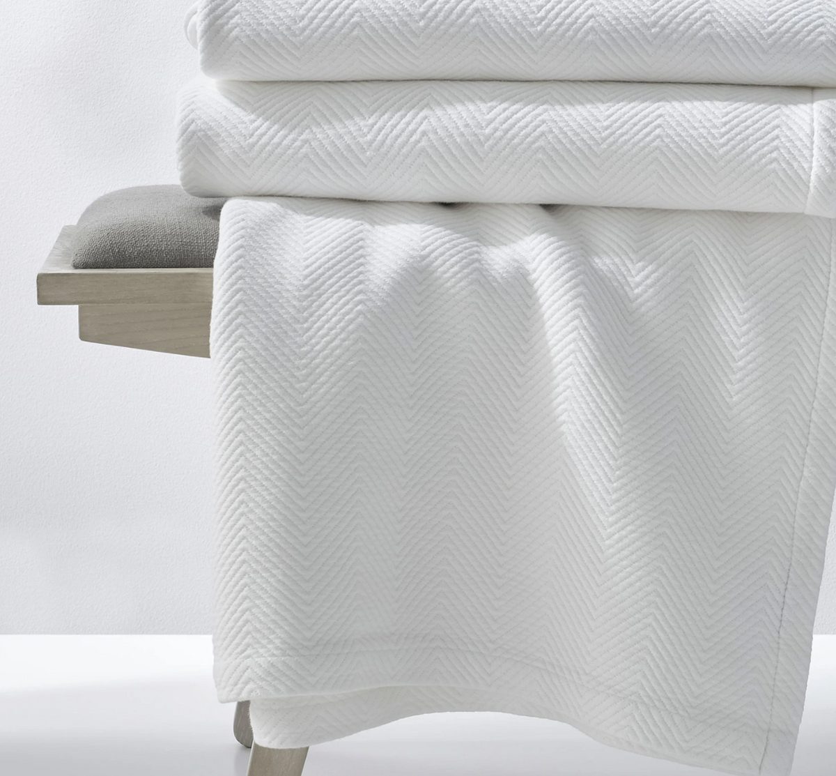 Folded white Espinho queen coverlet draped over a bench showcasing subtle chevron quilted texture on a light background