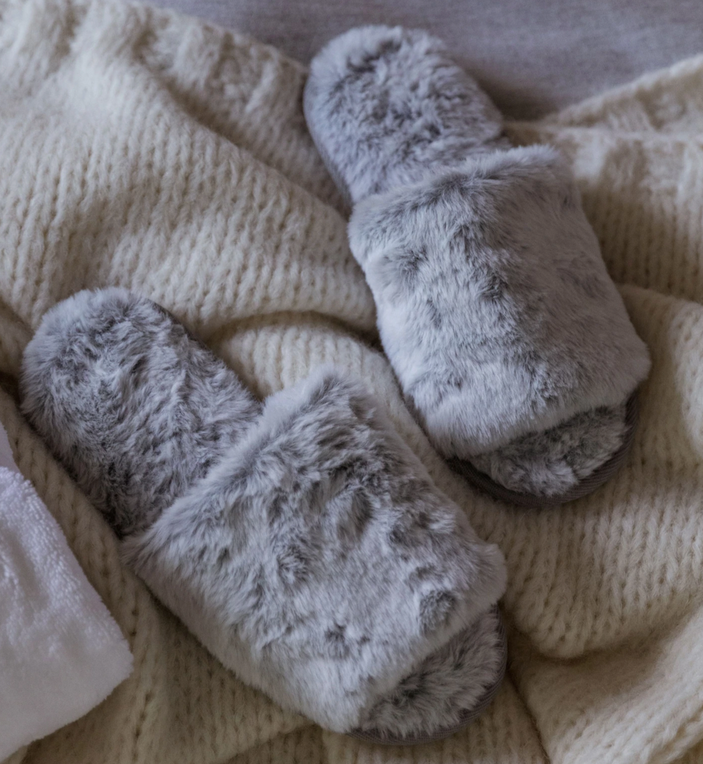 Top-down view of gray faux fur slippers showcasing plush faux-fur upper and cushioned footbed with cream knit blanket background