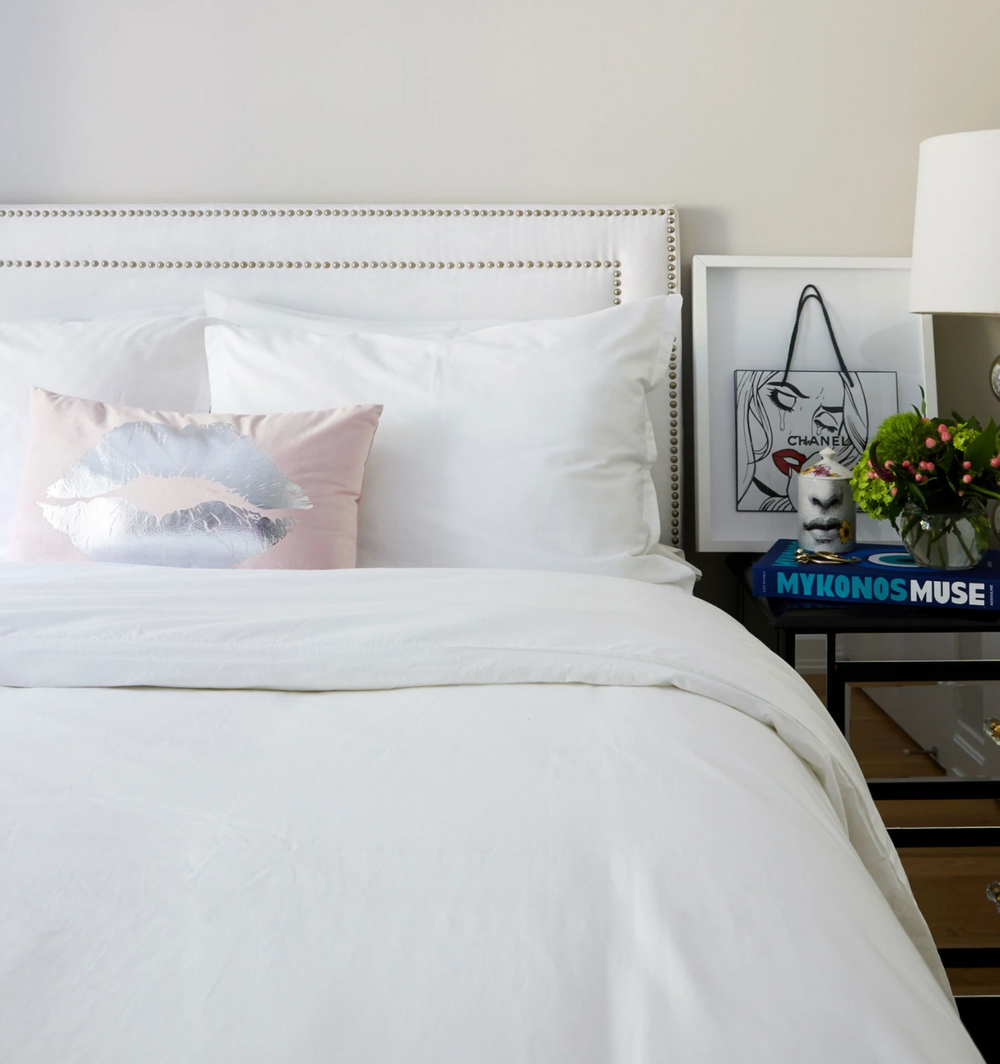 King bed styled with white Lorimer fitted sheet, pink metallic lip accent pillow, and nightstand with lamp and framed art
