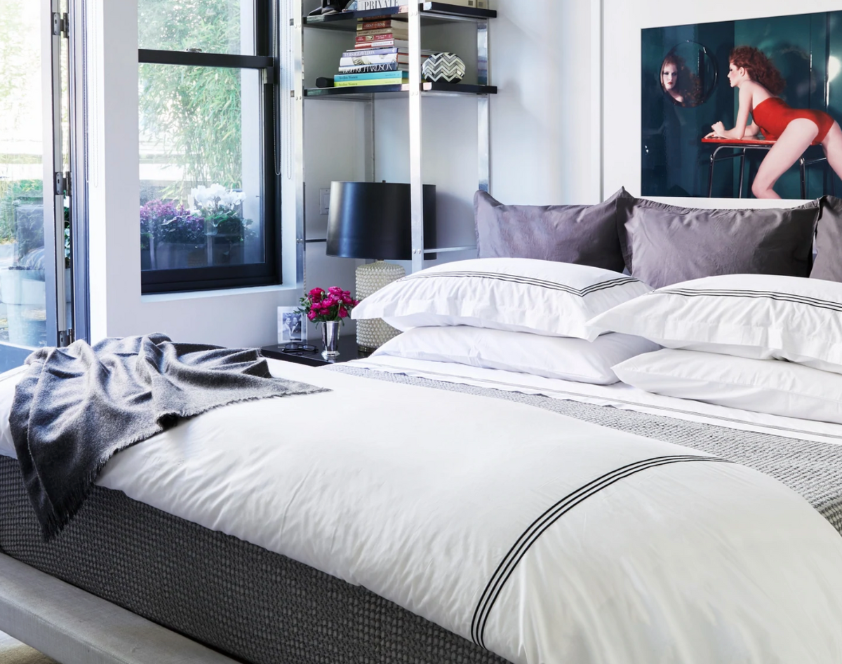 Angled view of Strada King flat sheet in white showing three-stripe black embroidery on a styled bed in a modern bedroom