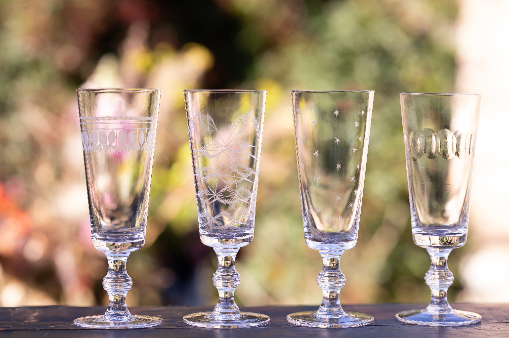 Front view of four crystal champagne flutes showcasing engraved oval patterns and short twisted stems on a blurred garden background
