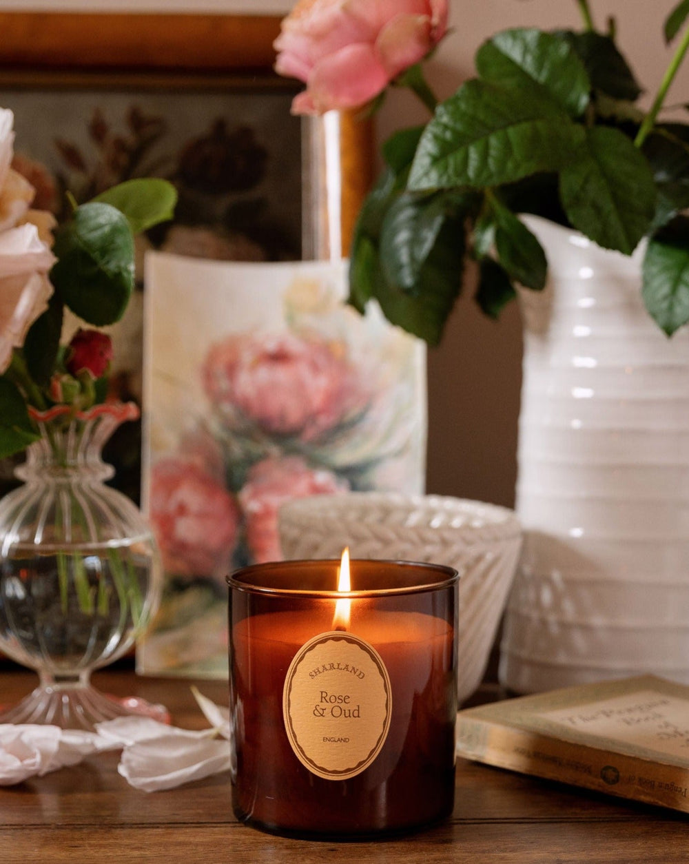Front view of Rose & Oud candle in amber glass jar with lit wick and paper label on wooden table with roses