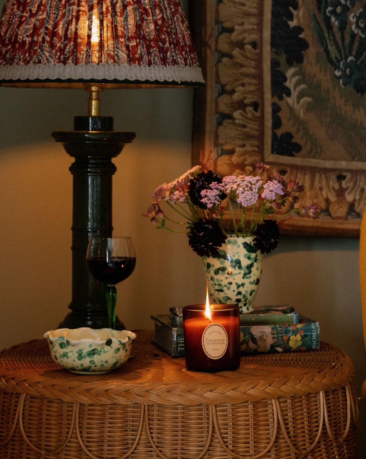 Close up of Pomegranate and Patchouli candle lit in amber glass jar showing flame and label on wicker table with lamp and flowers