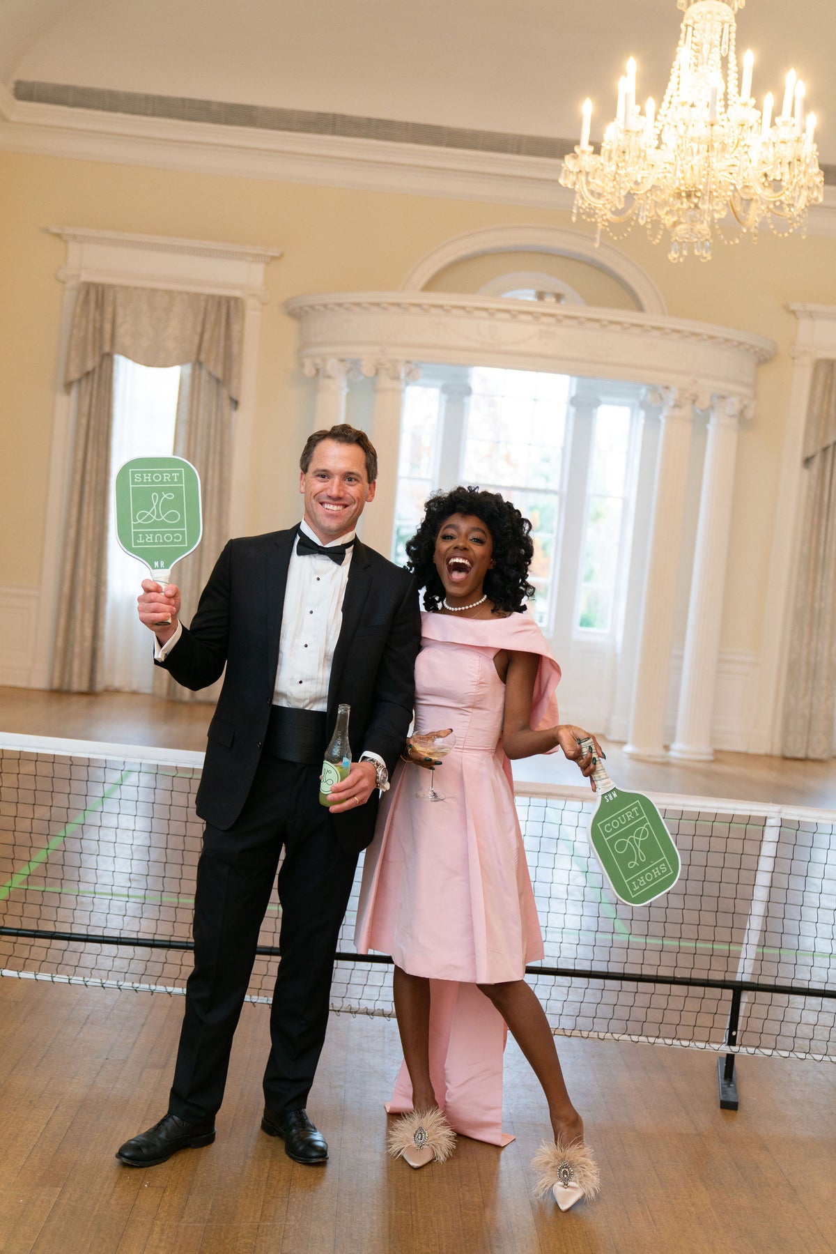 Man in tuxedo and woman in pink dress holding Short Court pickleball paddles and drinks beside an indoor net in a ballroom