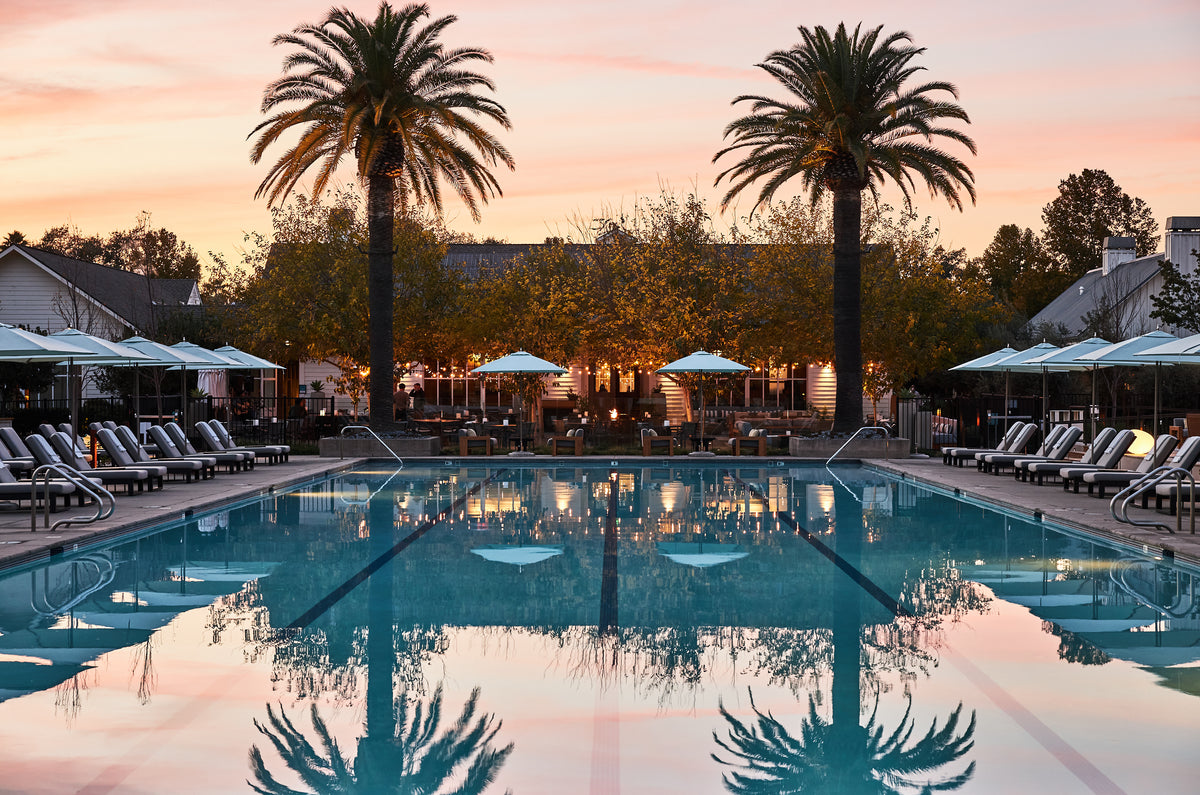 Resort pool at sunset with palm trees, lounge chairs, and umbrellas reflecting in the water