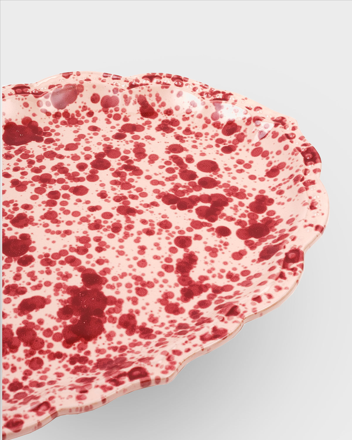Close-up view of Speckled Serving Plate in Pink showcasing red splatter glaze and scalloped oval edge with white background