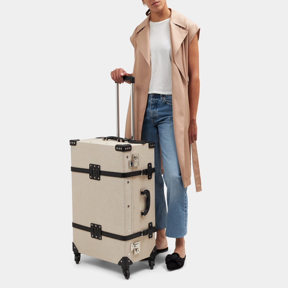 Front view of The Editor Check In Spinner showing ivory body, black straps, and extended trolley handle beside a model