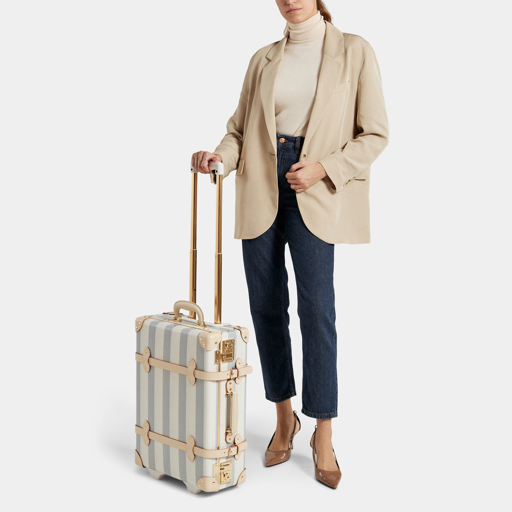 Three-quarter view of The Illustrator blue carryon with blue-and-white stripes, gold handle and tan leather straps on white background