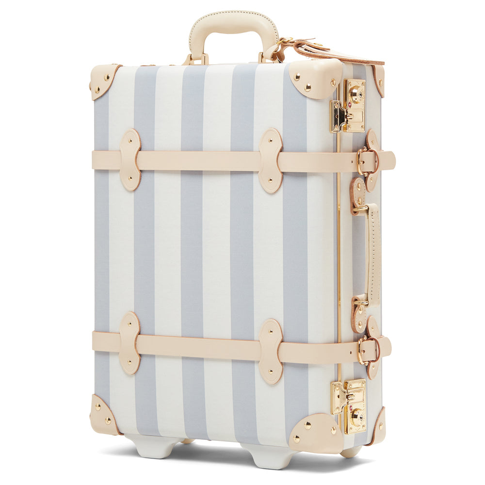 Three-quarter front view of The Illustrator Blue carry-on showcasing vertical blue and white stripes, beige leather straps and gold hardware on a white background