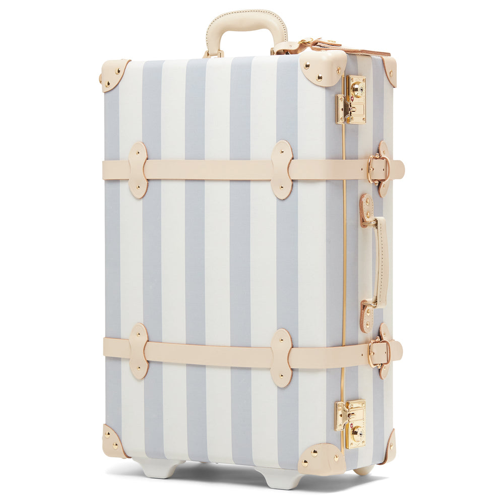 Three-quarter view of The Illustrator Blue Stowaway suitcase with blue-and-white vertical stripes, cream leather straps, and gold hardware on white background