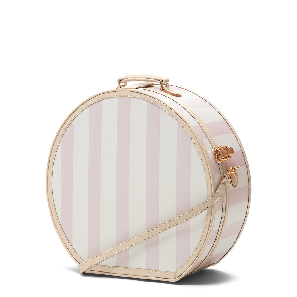 Three-quarter view of The Illustrator Deluxe pink hatbox showing vertical pink-and-white stripes, tan leather trim, top handle, and gold hardware on white background